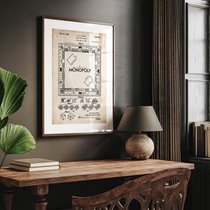 Op de afbeelding: Een ingelijste vintage Monopoly bordspel patent print. De print toont de lay-out van het spelbord, dobbelstenen en spelstukken. De lijst is donkerbruin van kleur en de print heeft een sepia-tint. Het woord "MONOPOLY" is prominent aanwezig.