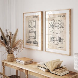 May include: Two framed vintage patent prints of a gaming console and controller. The prints are in wooden frames and feature detailed technical drawings on aged paper. The console print shows various views, while the controller print highlights its design.