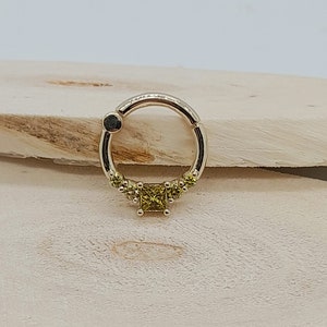 May include: A gold ring with a small square-shaped green gemstone in the center and smaller green gemstones around the center stone. The ring is on a wooden surface.