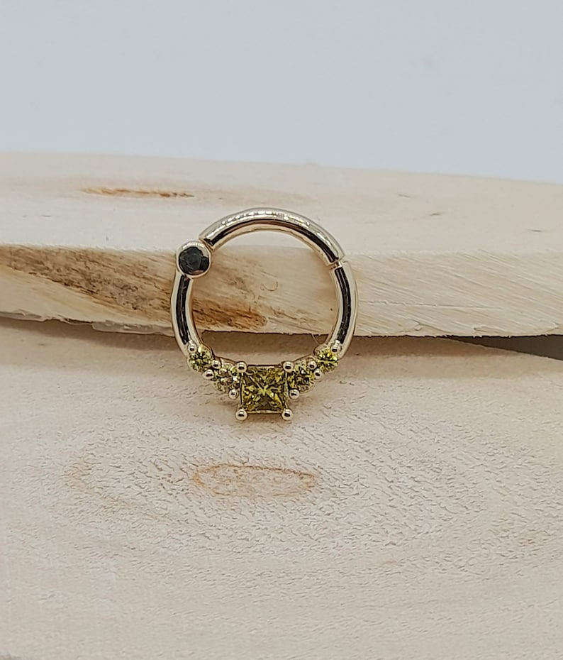 May include: A gold ring with a small square-shaped green gemstone in the center and smaller green gemstones around the center stone. The ring is on a wooden surface.