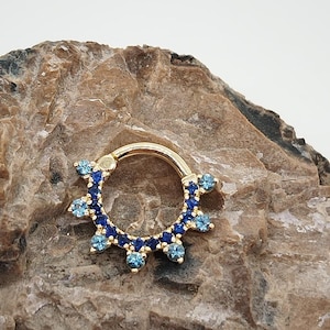 May include: A gold septum ring with a row of blue and light blue gemstones around the ring.