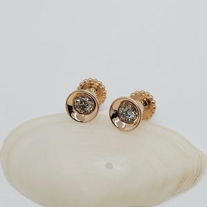 May include: A pair of gold stud earrings with a small round diamond in the center of each earring. The earrings are set in a circular bezel setting and have a textured edge.