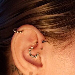 May include: A close-up of a person's ear with multiple piercings. The ear is pierced with a gold hoop earring, a gold ring with small diamonds, a black stud, and a gold stud. The ear is also pierced with a gold hoop earring with small diamonds.