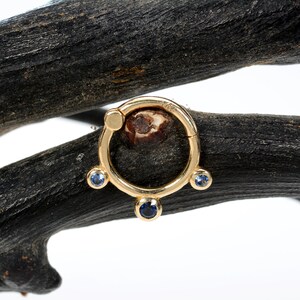Può includere: Orecchino a cerchio d'oro con tre pietre blu. L'orecchino è su una superficie di legno marrone scuro.