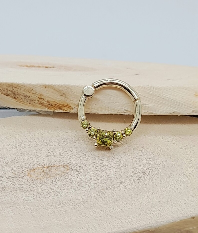 May include: A gold ring with a square-shaped green gemstone in the center and smaller green gemstones surrounding it.