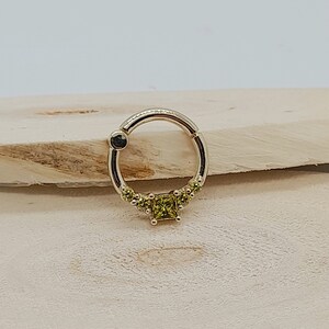 May include: A gold ring with a square-shaped green gemstone in the center and smaller green gemstones around the edge of the ring.