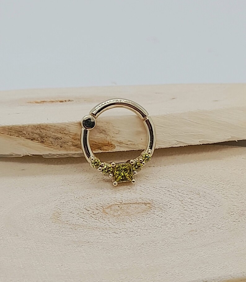 May include: A gold ring with a square-shaped green gemstone in the center and smaller green gemstones around the edge of the ring.