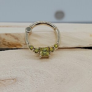 May include: A gold ring with a single large square-shaped green gemstone and smaller round green gemstones on either side of the large gemstone.