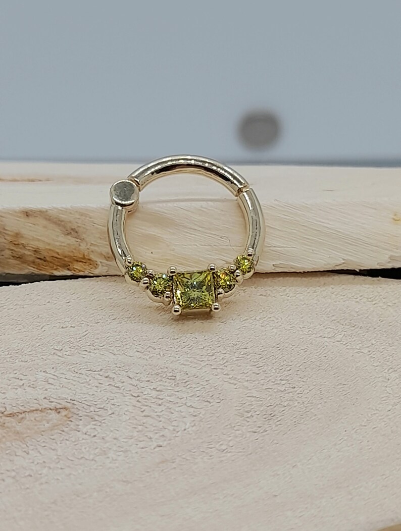May include: A gold ring with a single large square-shaped green gemstone and smaller round green gemstones on either side of the large gemstone.