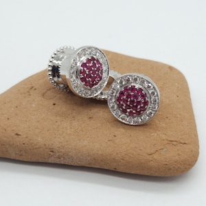 May include: A pair of silver earrings with a round red ruby stone surrounded by a halo of smaller white stones.