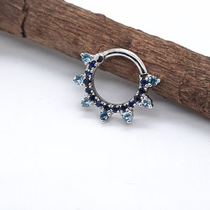 May include: A silver ring with a blue and light blue gem setting. The ring is set with small round gems that are arranged in a circle around the ring. The ring is a body piercing jewelry piece.