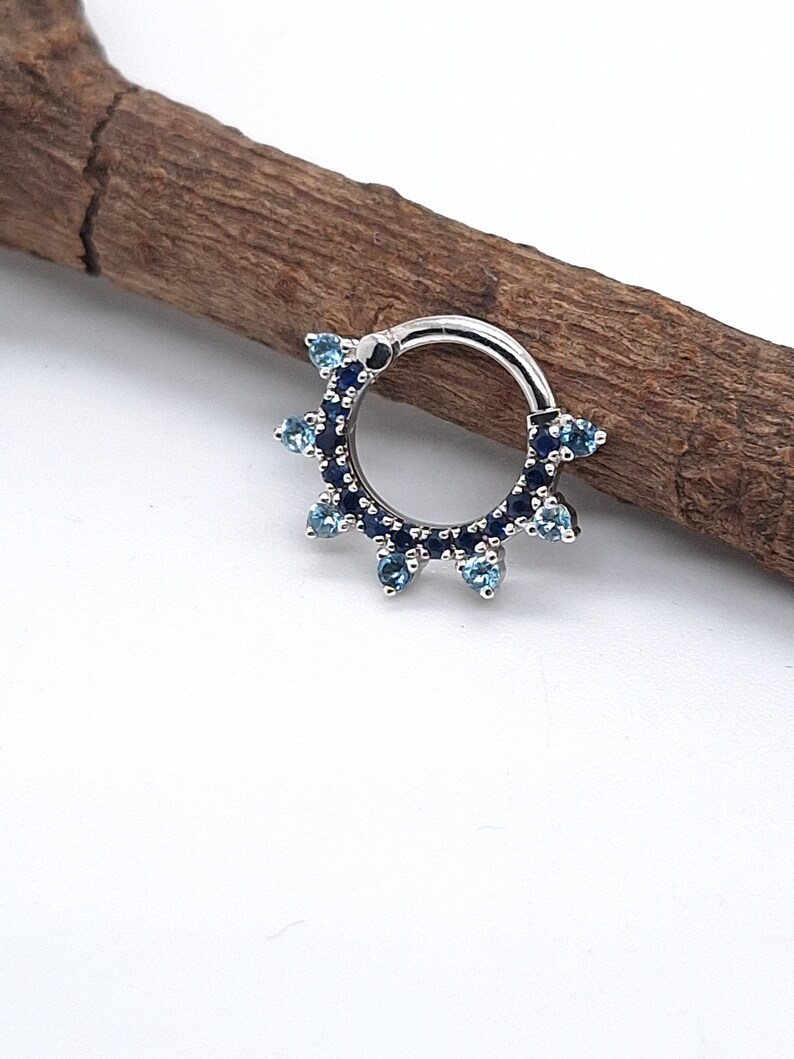 May include: A silver ring with a blue and light blue gem setting. The ring is set with small round gems that are arranged in a circle around the ring. The ring is a body piercing jewelry piece.