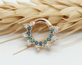 Puede incluir: Un anillo de clic dorado con un círculo de piedras preciosas azules y blancas. El anillo está diseñado con una fila de piedras preciosas puntiagudas alrededor del borde.