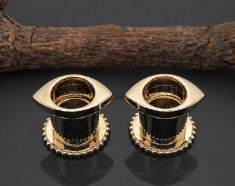 May include: Two gold-colored, eye-shaped ear gauges. Each gauge has a cylindrical body with a flared top and a beaded edge. The gauges are displayed on a dark surface with a piece of wood in the background.