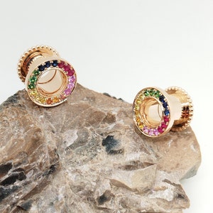 May include: A pair of gold-toned ear gauges with a rainbow of colorful gemstones set around the outer edge. The gauges are on a rock surface.