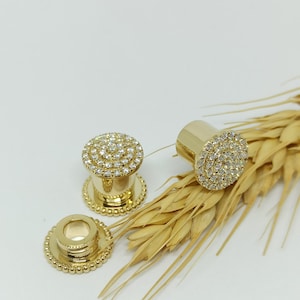 May include: A pair of gold-toned ear gauges with a circular design featuring a ring of small, clear stones. The gauges are displayed on a background of wheat stalks.