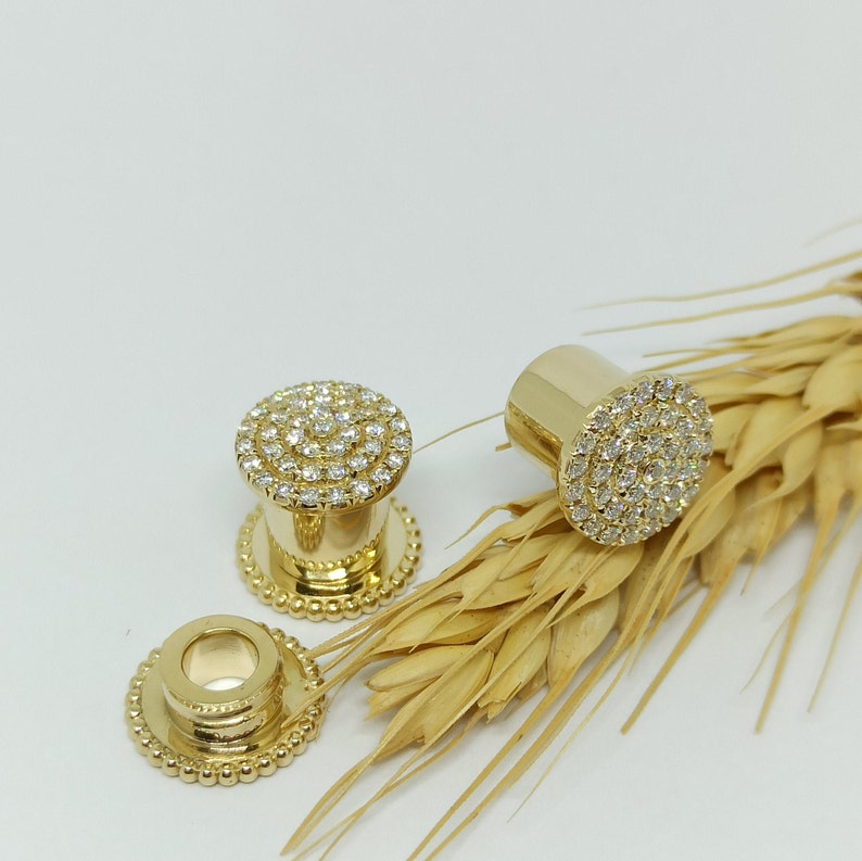 May include: A pair of gold-toned ear gauges with a circular design featuring a ring of small, clear stones. The gauges are displayed on a background of wheat stalks.