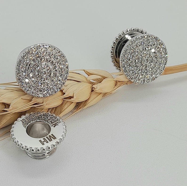 May include: Two silver tone earrings with a round, clear crystal design. The earrings are on a light brown background.