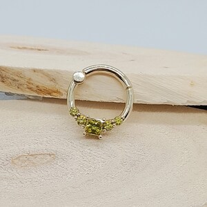 May include: A gold septum ring with a cluster of small, square-shaped, light green gemstones. The ring is open and has a small bead at the end of the opening.