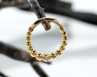 May include: A gold-colored beaded hoop earring with a small, clear gemstone. The earring is suspended from a dark, thin branch against a white background. The earring is circular and features small, evenly spaced beads.