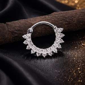 Op de afbeelding: Een zilverkleurige septumring met een zonnesterontwerp. De ring is versierd met talrijke kleine, heldere edelstenen, die een sprankelend effect creëren. De sieraden zijn geplaatst tegen een donkere achtergrond.