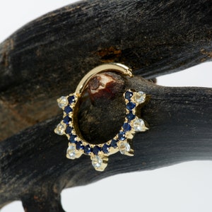May include: A gold hoop earring with a row of blue and white gemstones. The hoop is set with small, round gemstones that are arranged in a circle. The gemstones are a mix of blue and white, and they are set in a gold setting.