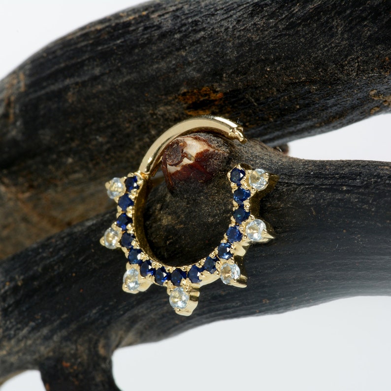 May include: A gold hoop earring with a row of blue and white gemstones. The hoop is set with small, round gemstones that are arranged in a circle. The gemstones are a mix of blue and white, and they are set in a gold setting.