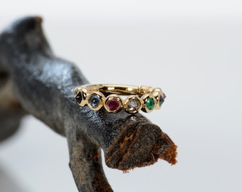 May include: A gold ring with a series of colorful gemstones, including blue, red, and green stones. The ring is set on a dark, textured branch against a white background. The gemstones are round and evenly spaced.