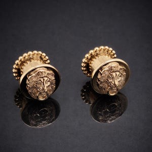 Op de afbeelding: Een paar goudkleurige oordopjes met een gedetailleerd leeuwenkopontwerp. De oordopjes hebben een afgeronde basis met een kralenrand en een plat, cirkelvormig vlak met de leeuwenkop. De achtergrond is donker.
