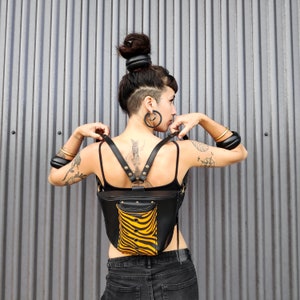 Puede incluir: Mochila de cuero negro con un diseño único. La mochila presenta un pequeño bolsillo frontal con un estampado de cebra amarillo y negro. Las correas son ajustables y la bolsa tiene una forma curva. El fondo es una pared de metal corrugado.