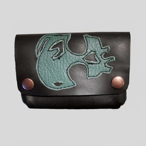 May include: Black leather pouch featuring a teal emblem stitched on the front. The pouch has a flap closure secured by two copper rivets. The design is likely for a card game. The pouch is approximately 10 cm wide.
