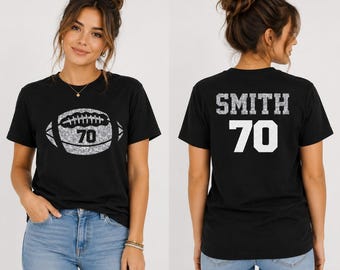 Könnte beinhalten: Ein schwarzes Kurzarm-T-Shirt mit einer silbernen Glitzer-Grafik eines Footballs und der Zahl 70 auf der Vorderseite. Auf der Rückseite des T-Shirts steht der Name SMITH in silbernen Glitzerbuchstaben über der großen weißen Zahl 70.