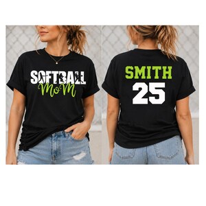 May include: A black short-sleeved t-shirt featuring the white text SOFTBALL with a silhouette of a player and the green script text MOM on the front. The back of the shirt displays the green text SMITH above the large white number 25.