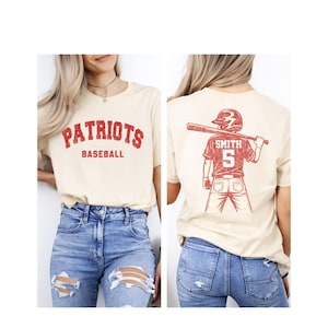 May include: Cream-coloured t-shirt with the words "PATRIOTS BASEBALL" in red. The back of the shirt has a red graphic of a baseball player with the name "SMITH" and the number "5". Worn with blue jeans.