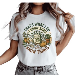 May include: White t-shirt with a circular graphic featuring gardening tools, plants, and the text "That's What I Do, I Grow Stuff and Know Things." The design includes a watering can, gloves, and potted plants. The shirt is worn with denim shorts.