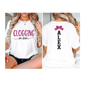 Puede incluir: Camiseta blanca con el texto "CLOGGING mom" en letras brillantes rosas en la parte delantera. La parte trasera de la camiseta presenta el nombre "ALEX" en letras negras con un lazo brillante rosa encima. La camiseta es de estilo casual.
