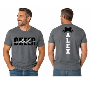 May include: A charcoal gray short-sleeve t-shirt featuring the word CHEER in bold black letters with the word dad in white script across the front. The back displays the name ALEX in white vertical block letters topped with a black bow graphic.