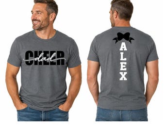 Könnte beinhalten: Ein anthrazitfarbenes Kurzarm-T-Shirt mit dem Wort CHEER in fetten schwarzen Buchstaben und dem Wort dad in weißer Schreibschrift auf der Vorderseite. Die Rückseite zeigt den Namen ALEX in vertikalen weißen Blockbuchstaben mit einer schwarzen Schleife.