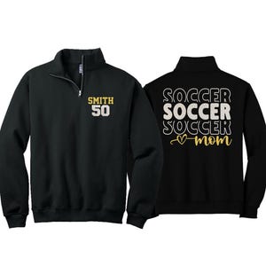 May include: Black quarter-zip sweatshirt with gold lettering. The front displays "SMITH 50", while the back features "SOCCER SOCCER SOCCER mom" in white and gold. A casual, sporty top.