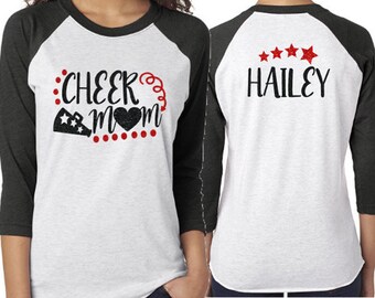May include: A white and black baseball style shirt with the text "Cheer Mom" and a heart on the front. The back of the shirt has the text "Hailey" with five red stars above it.