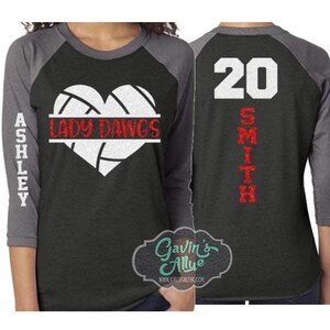 May include: A black and grey baseball t-shirt with a white volleyball in a red heart shape. The text "Lady Dawgs" is written on a red banner across the volleyball. The text "Ashley" is written vertically on the left sleeve and the text "Smith 20" is written vertically on the right sleeve.