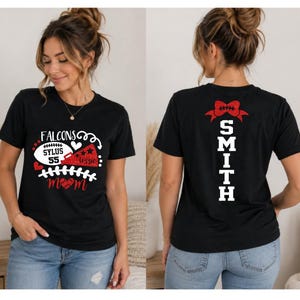 Op de afbeelding: Een zwart t-shirt met korte mouwen en een wit-rode afbeelding op de voorkant met de tekst "FALCONS", "SYLUS 55" en "MOM", samen met een football en een megafoon. De achterkant toont de naam "SMITH" in witte letters met een rode strik.