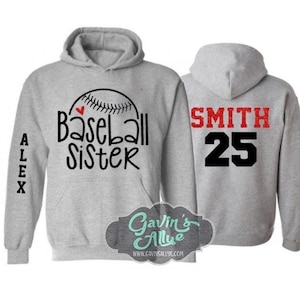 May include: Gray hooded sweatshirt with the text "Baseball Sister" and a red heart on the front. The left sleeve has the name "ALEX" printed vertically. The back of the sweatshirt has the text "SMITH 25" in red and black.