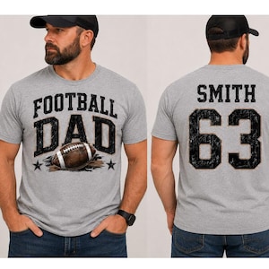 May include: A light gray short-sleeved t-shirt featuring a graphic print. The front displays the text FOOTBALL DAD with a football illustration. The back shows the name SMITH and the number 63 in a distressed black font with a dark outline.