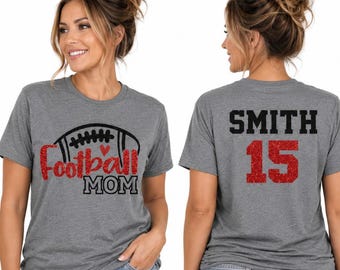 Könnte beinhalten: Ein graues, meliertes Kurzarm-T-Shirt mit einer Vorderseite, die einen schwarzen Football-Umriss und den roten Glitzertext Football MOM zeigt, sowie einer Rückseite mit dem schwarzen Text SMITH über der roten Glitzerzahl 15.