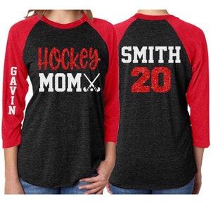 May include: Black and red baseball style shirt with the text "Hockey Mom" and hockey sticks on the front and the text "SMITH 20" on the back.