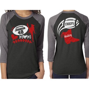 May include: Two black and grey baseball shirts with red glitter designs. The front shirt has a football, a cheerleader, a heart, and the text "Johnson 48 Mom". The back shirt has a football with the text "Chiefs" and a boot with the text "Hailey".