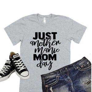 May include: A gray t-shirt with black text that reads "JUST another manic MOM day". The shirt is paired with black sneakers and blue denim shorts.