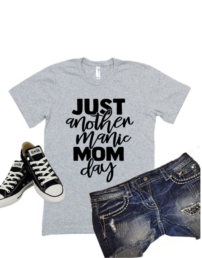 May include: A gray t-shirt with black text that reads "JUST another manic MOM day". The shirt is paired with black sneakers and blue denim shorts.