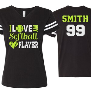 Op de afbeelding: Twee zwarte softbaltruien met witte strepen op de mouwen. Een trui heeft de tekst "I LOVE MY Softball PLAYER" met een groene softbalafbeelding en een hartafbeelding. De andere trui heeft de tekst "SMITH 99" in wit.
